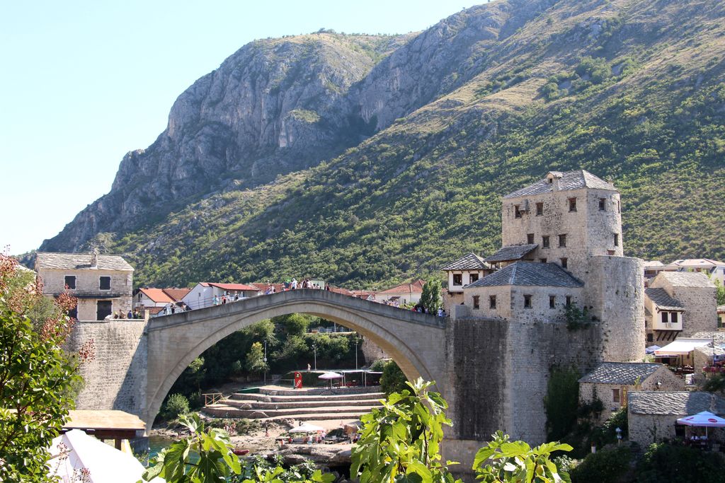 A single pale stone arch with a steep deck spans a blue-green river between masonry towers, town buildings and mountains.
