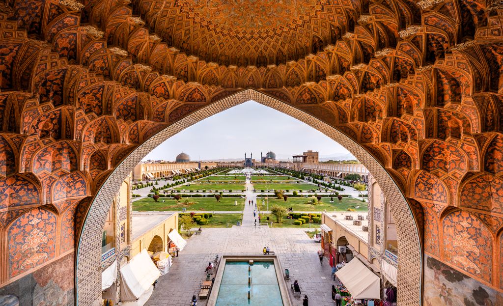 A richly painted vaulted arch frames a long green square with paths, water, people and distant domed buildings.