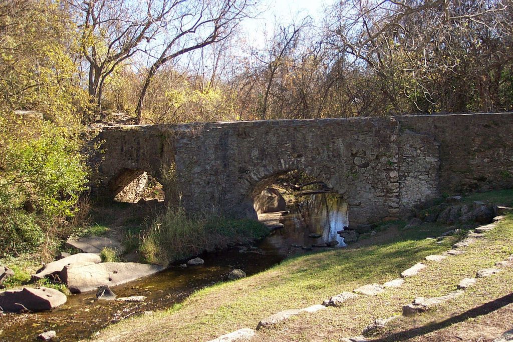Two low stone arches carry a water channel across a creek amid trees and grass.