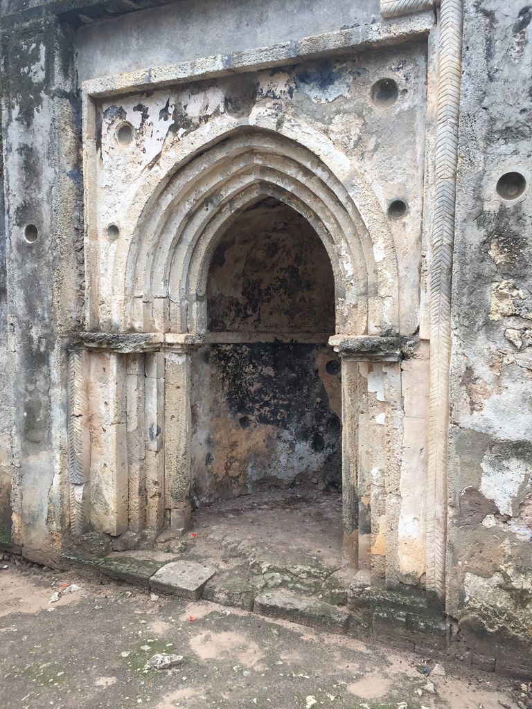 A tall pointed doorway with several recessed arches and carved vertical bands stands in a weathered pale masonry wall.
