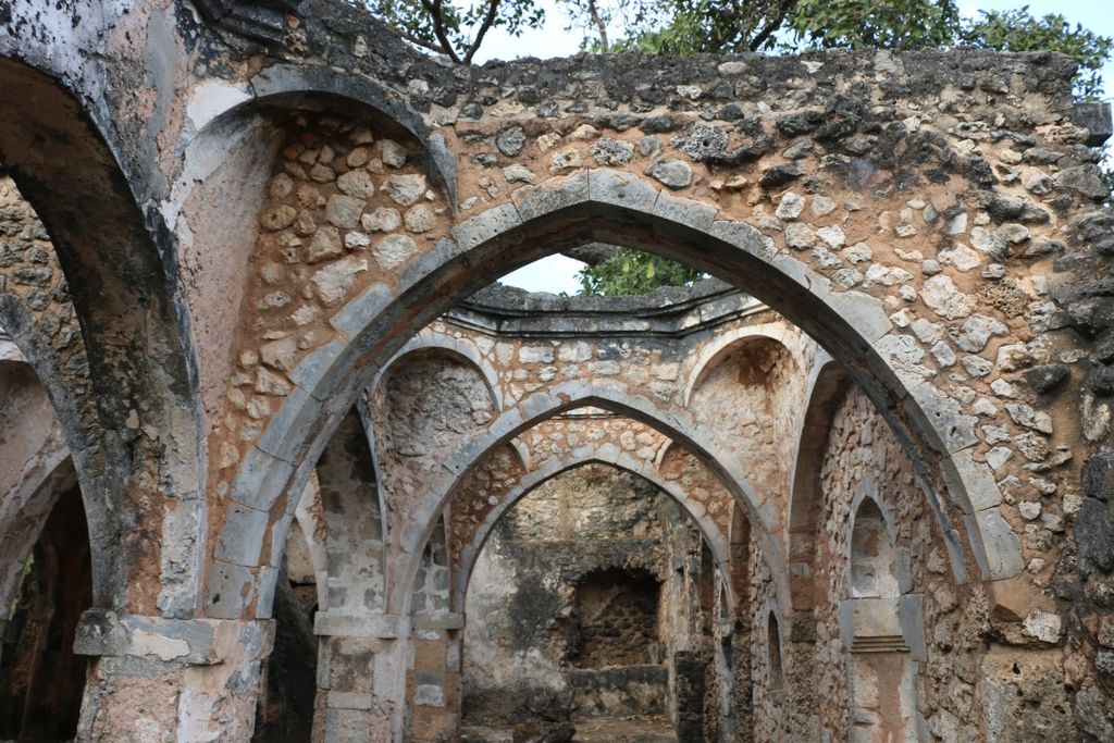 Successive pointed arches and wall niches cross the roofless interior of a weathered coral-stone mosque ruin beneath trees.