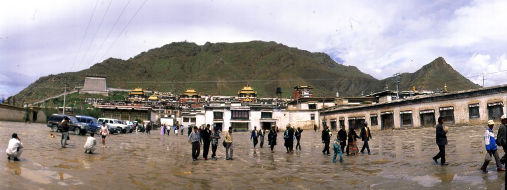 A wide paved forecourt with scattered people stretches before long white monastery ranges and coloured roofs beneath a green-brown hill.