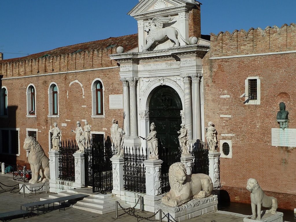 An elaborate white stone arched gate with a winged lion is set in a long crenellated brick wall behind iron gates and several large stone statues.