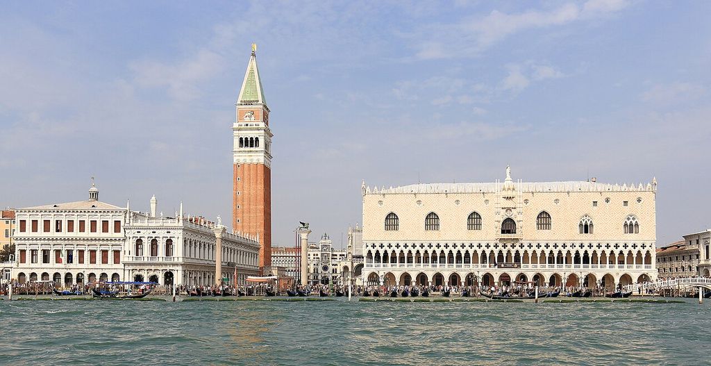 Across choppy lagoon water, the pale Doge's Palace has two open arcaded levels beneath a patterned upper wall, beside a tall brick campanile and classical civic buildings.