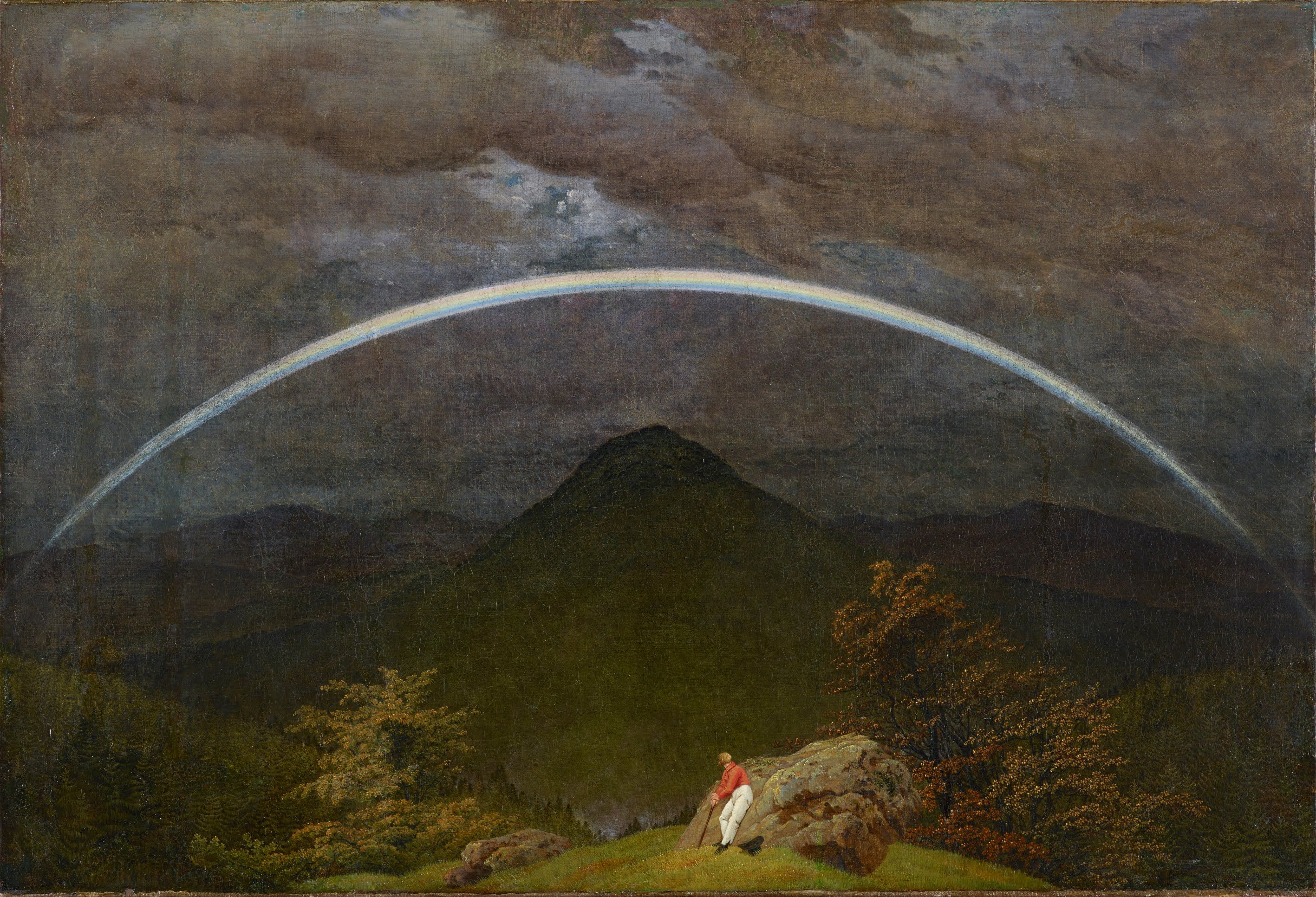 Friedrich's Mountain Landscape with Rainbow shows a lone man resting on a rocky foreground beneath a pale rainbow spanning a dark mountain valley.