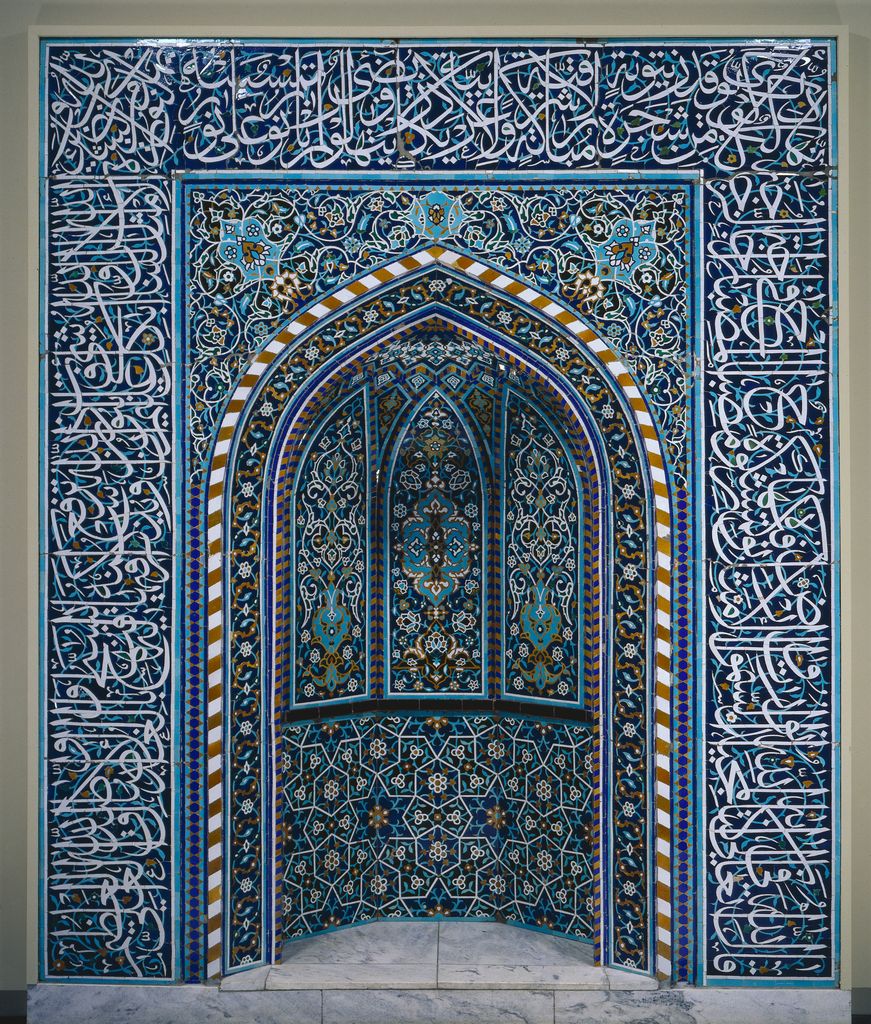 A pointed ceramic mosaic prayer niche combines white Arabic calligraphy with turquoise, cobalt, green, gold and dark fields.