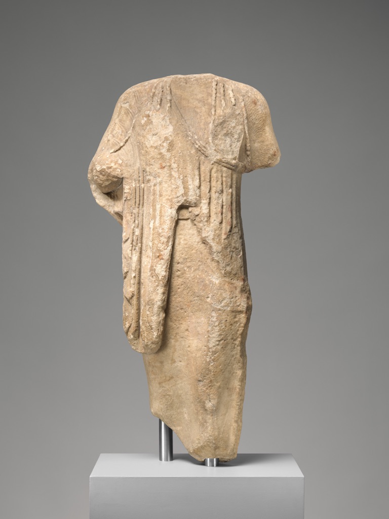 A headless marble kore stands on a mount with carved drapery folds, worn areas and rough broken surfaces visible across the stone.