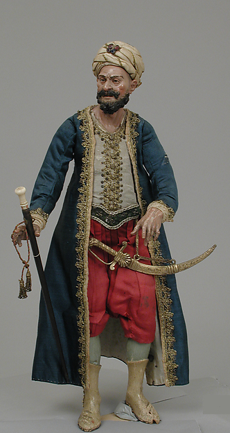 Dressed mixed-media figure with a terracotta head, wooden limbs, wire body and elaborate textiles.